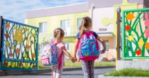 Two school-aged children hold hands and walk towards a colorful school.